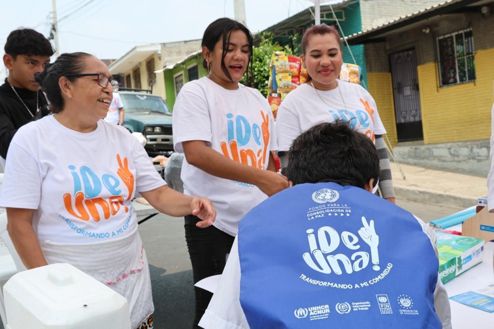 Three women in white tshirts stand at a desk where another person wearing a blue jacket is sitting. Their shirts are marked with the logo of the De Una programme.