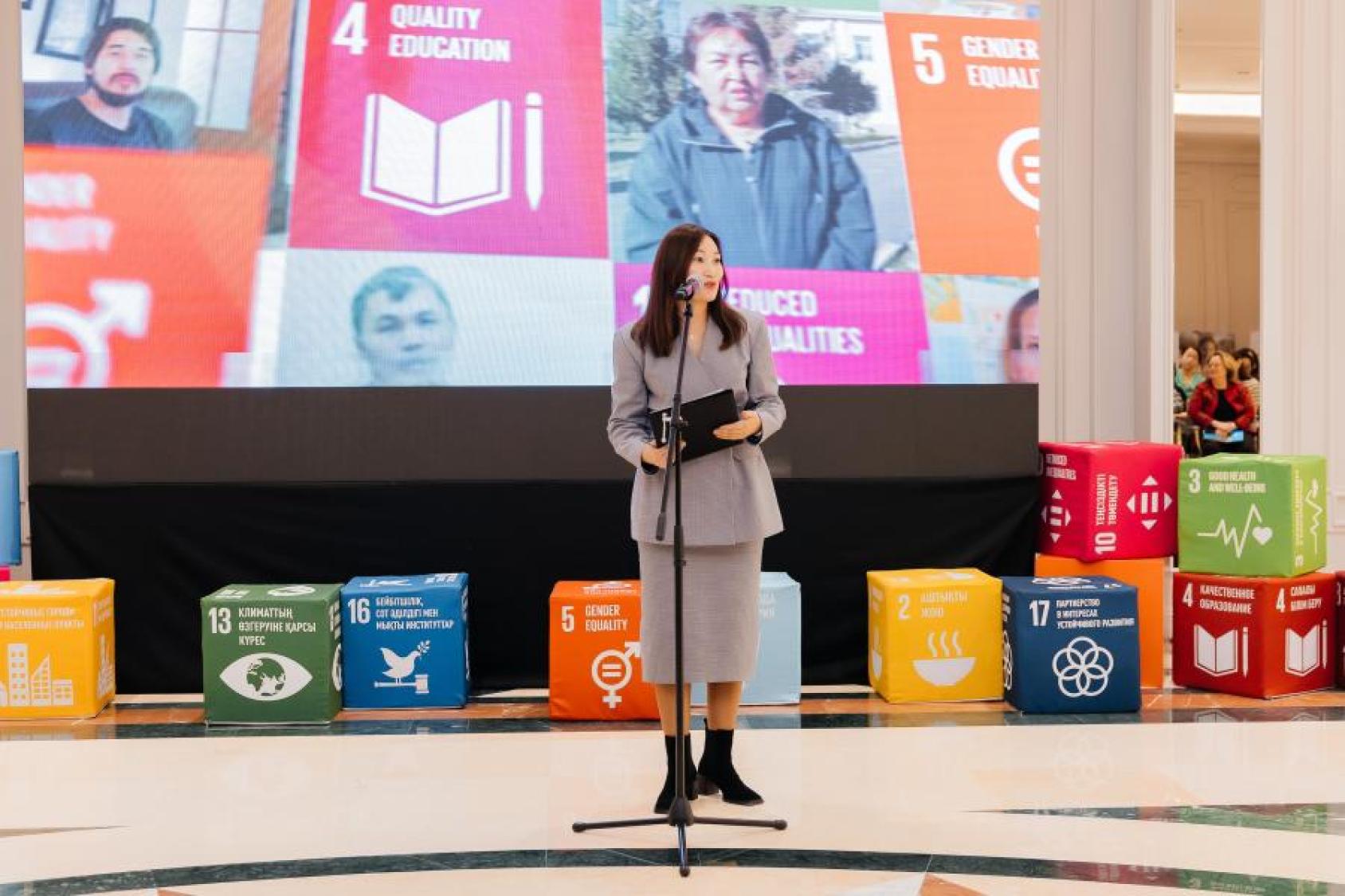 A woman in a grey formal suit speaks into a microphone in front of a big screen with symbols of the sustainable development goals