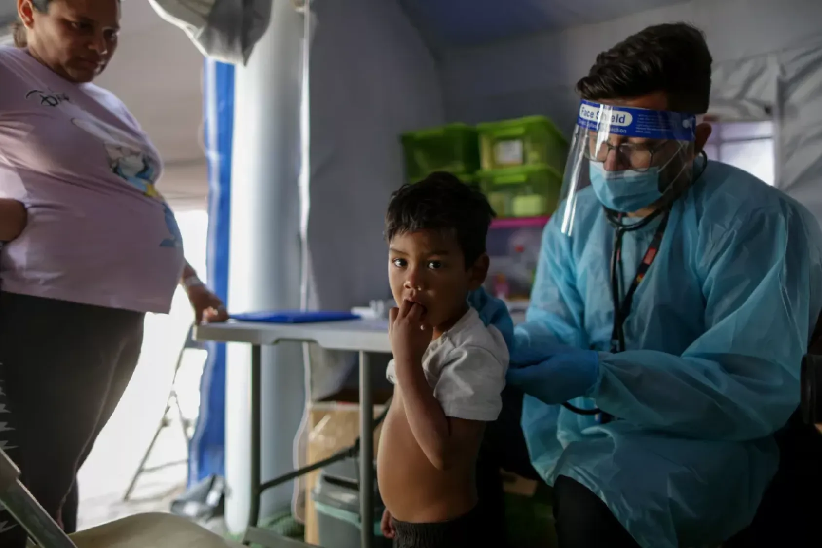 A doctor in a blue coat, mask and shield treats a young child as his mother looks on