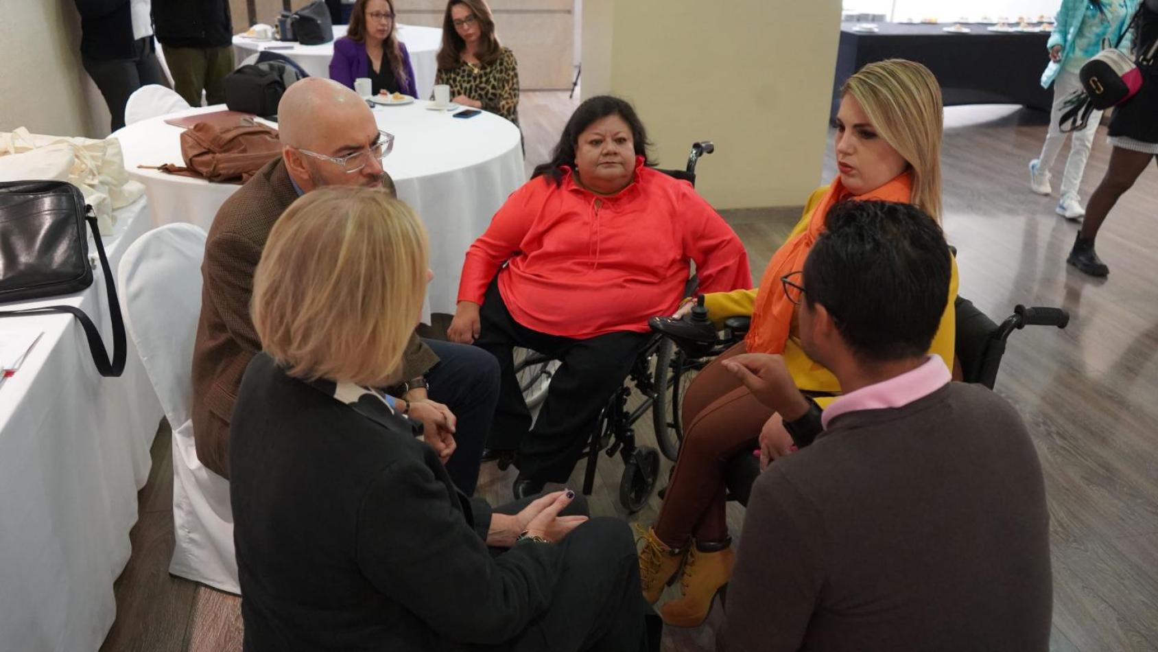 A group of five people gather and talk, the prominent speaker is a woman in an orange shirt in a wheelchair.