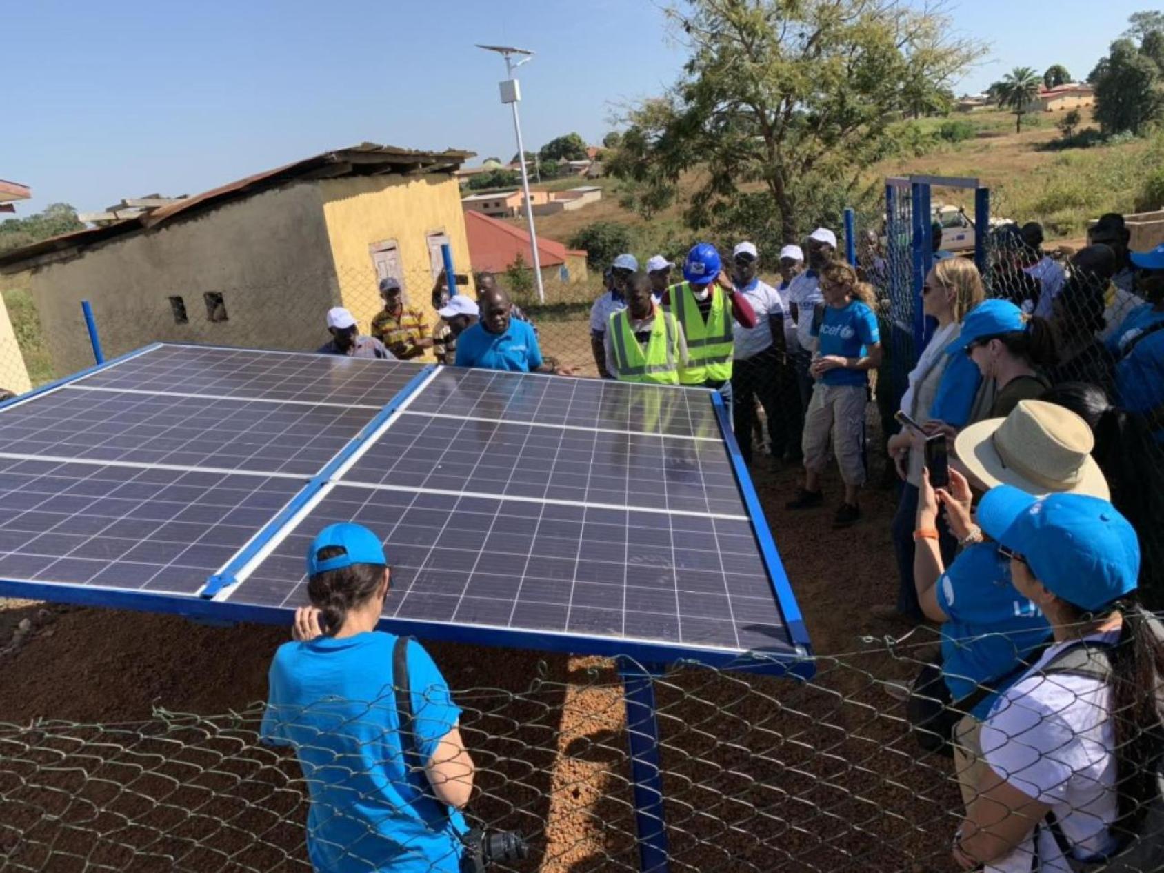 A group of people in blue shirts and hats gather around a silver solar panel in Guinea