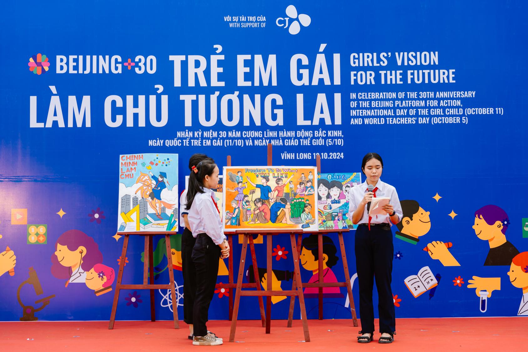 Two girls in school uniforms speak into a microphone on a stage, as they present their art work on an easel at an event commemorating gender equality.