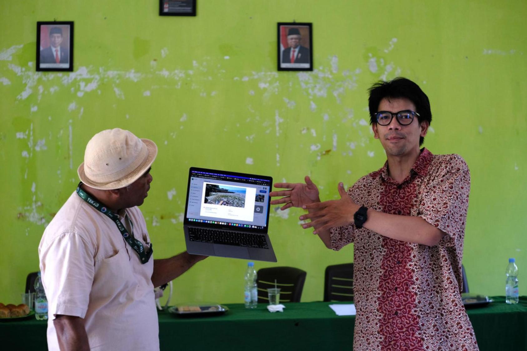 A man in a batik red shirt gesticulates as another man holds up a laptop with images on it