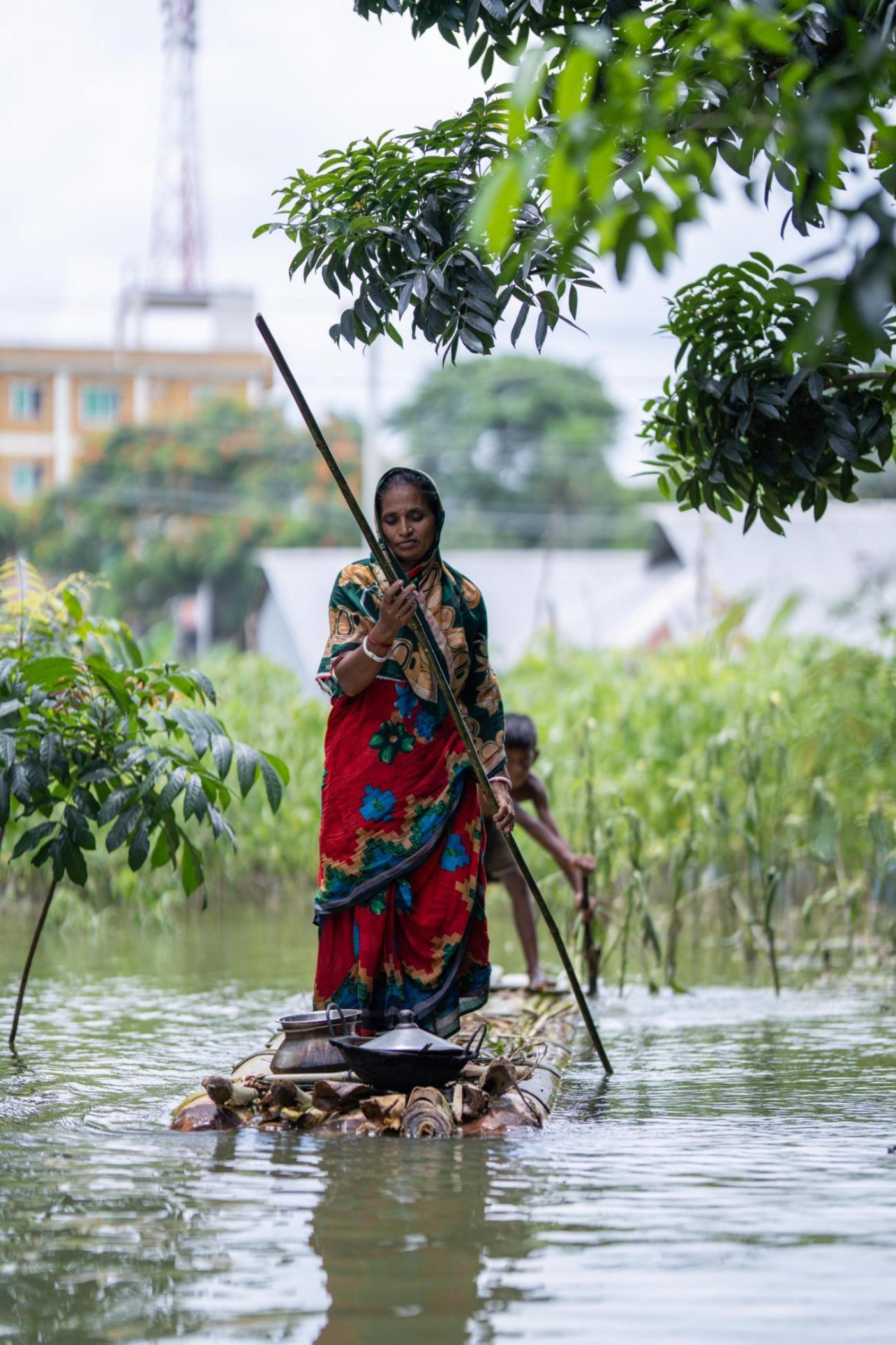 A woman in a red saree floats on a steel board using a long stick. Water surrounds her everywhere.