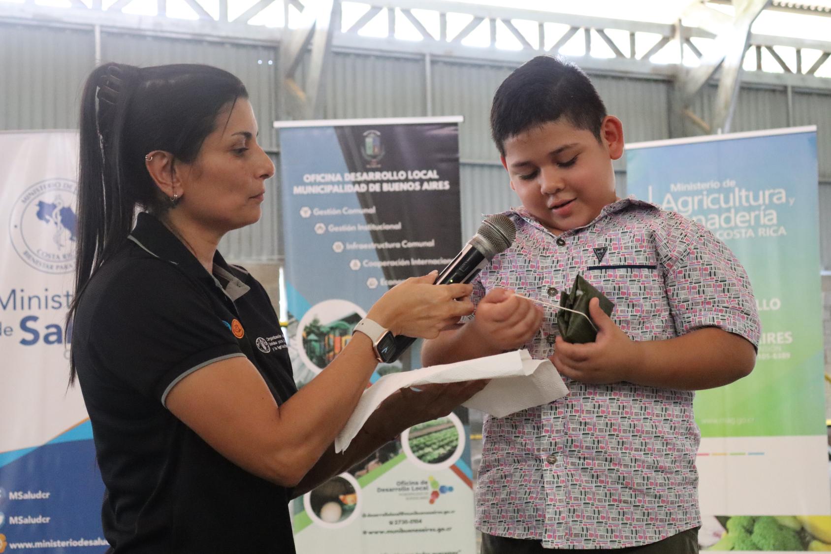 A woman in a black shirt holds up a microphone for a young boy dressed in a grey shirt who appears to be unwrapping a tamale.