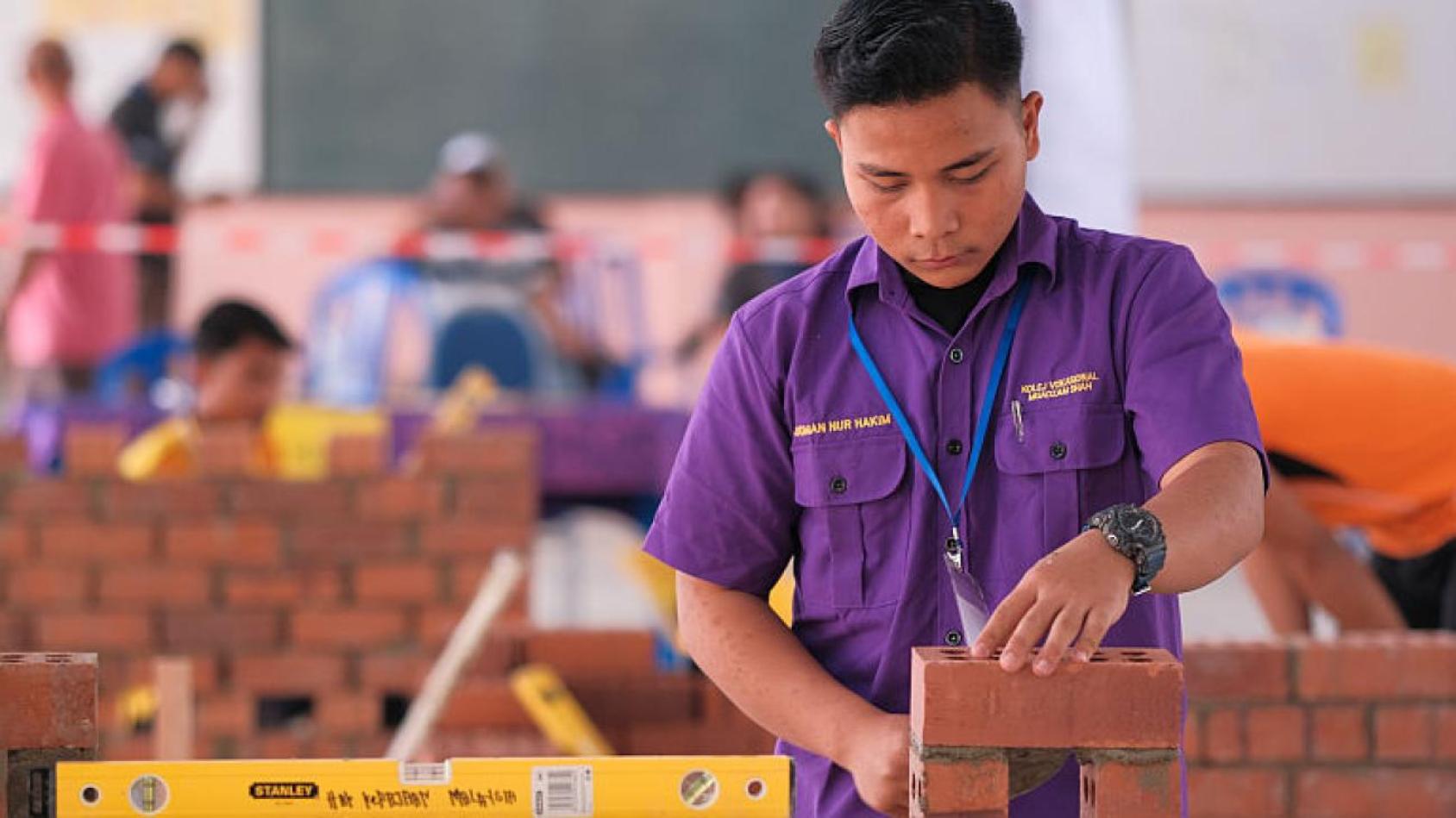 A man in a purple shirt tests out construction materials, laying bricks one after another.