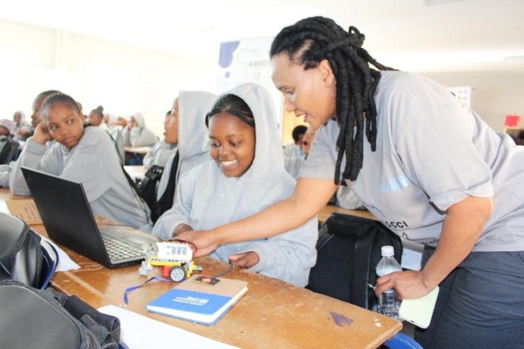 A woman, a teacher dressed in grey, peers over two young girls working on a computer in a classroom