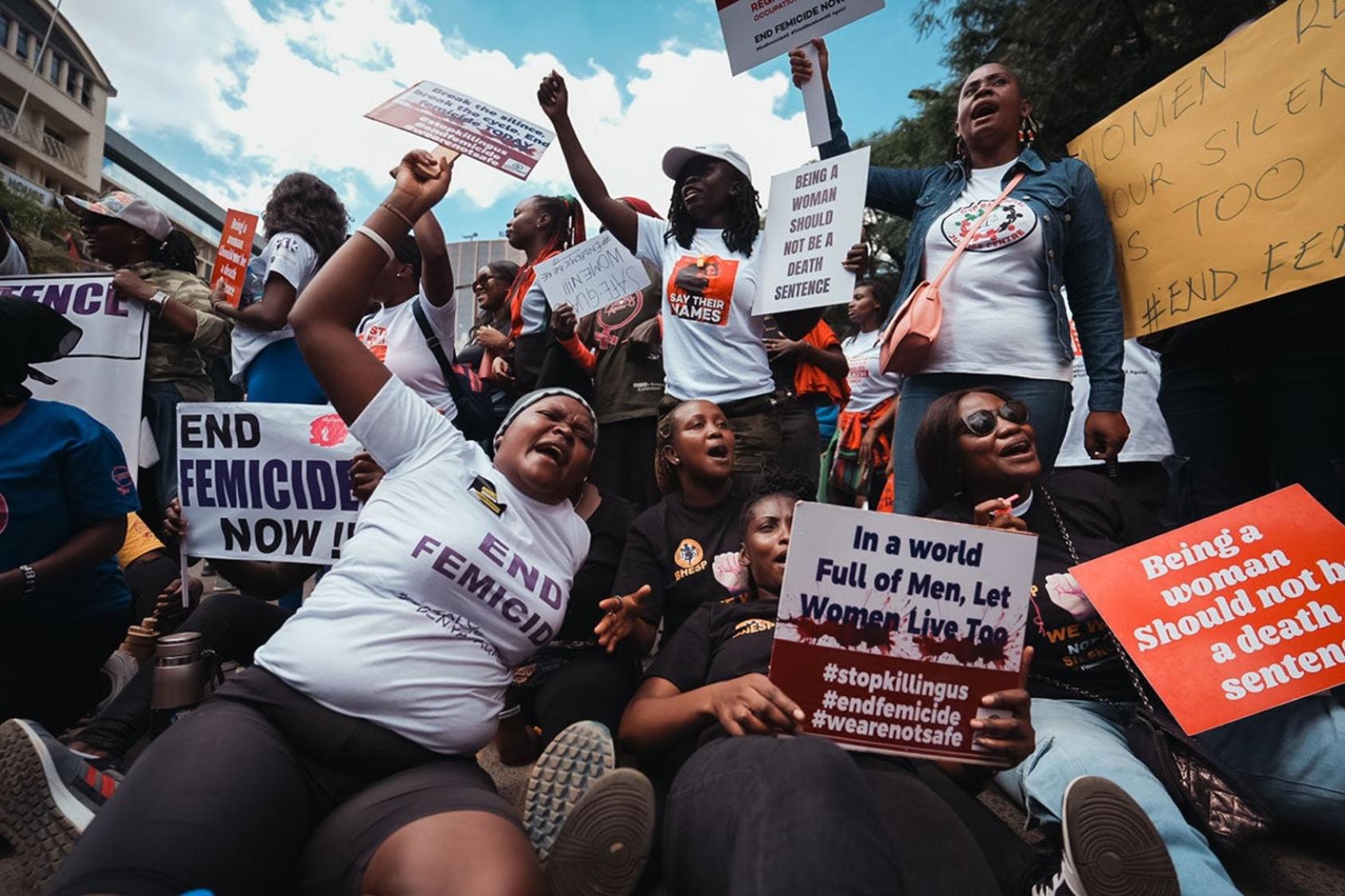 A group of women stand with posters and slogans, raising their fists in the air in protest.