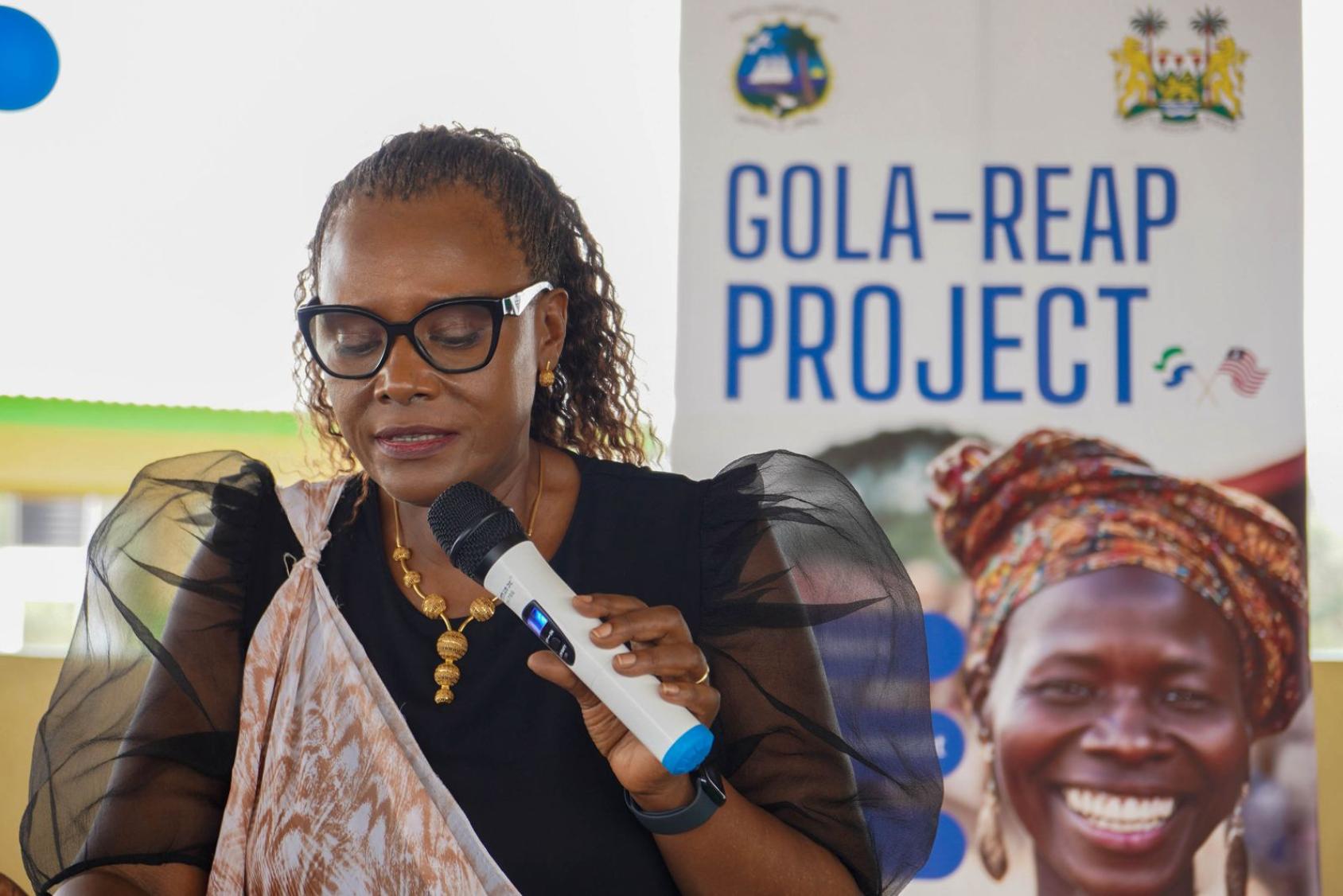 A woman in a black shirt and printed scarf speaks into a microphone at a podium. Behind her is a banner that says "GOLA REAP Project"
