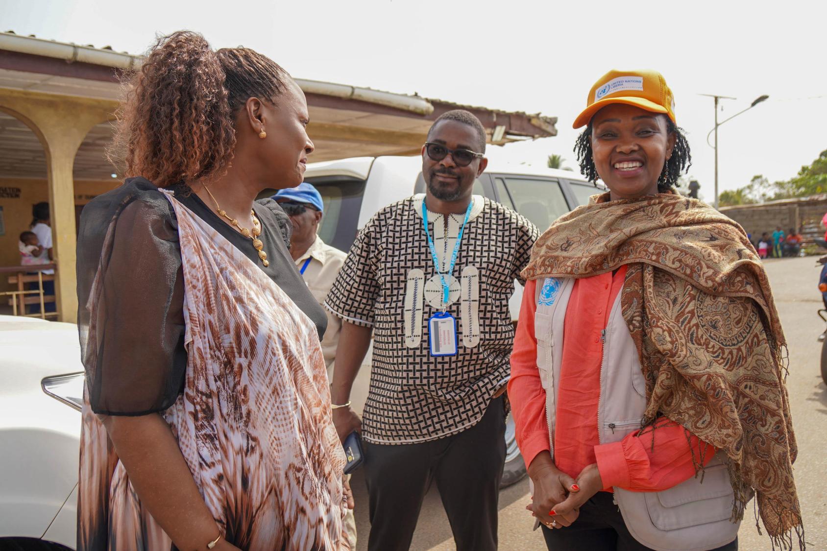 Two women, wearing colourful clothes and hats, the UN Resident Coordinators smile and talk to each other while a man watches on