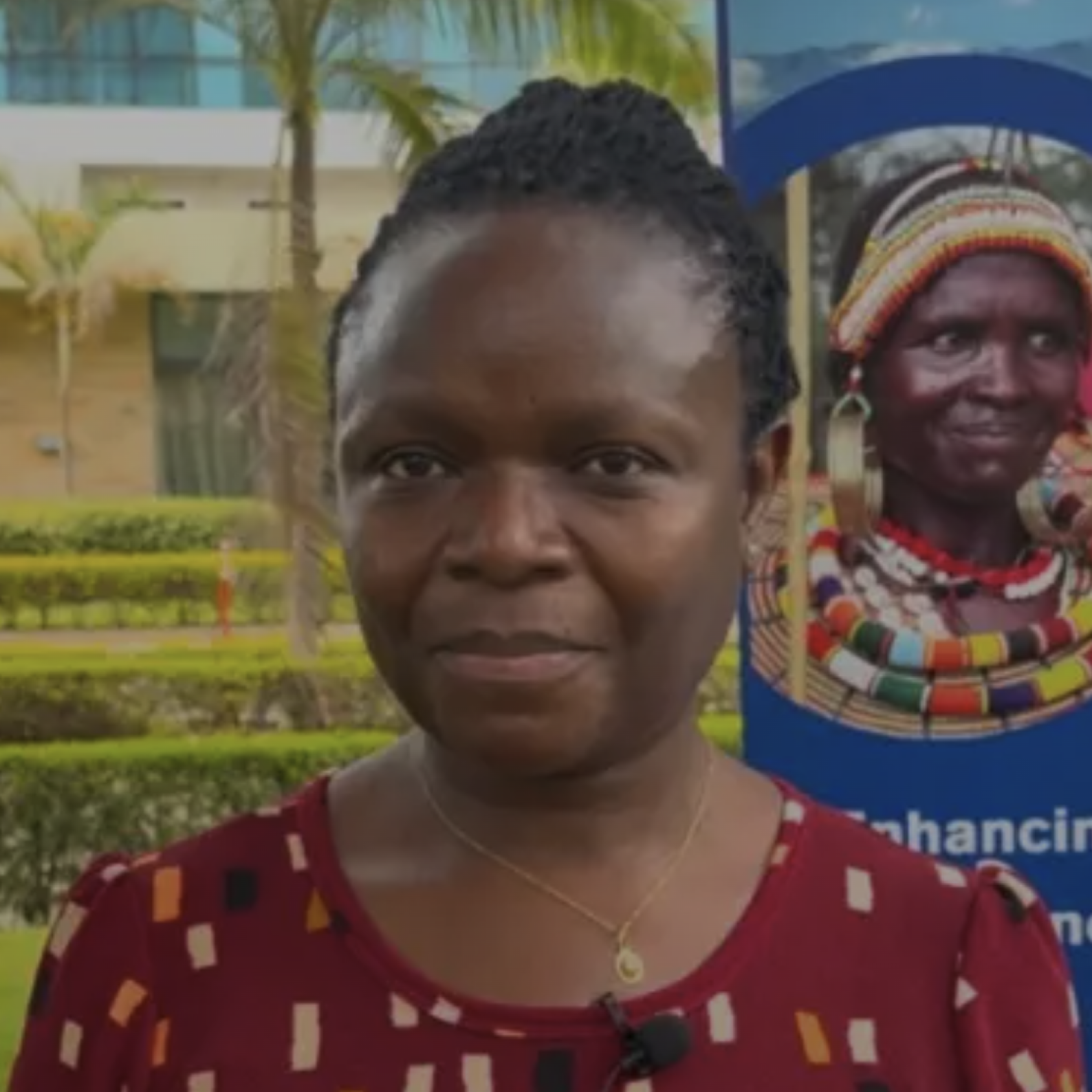 A woman stands in front of a building surrounded by grass and trees.
