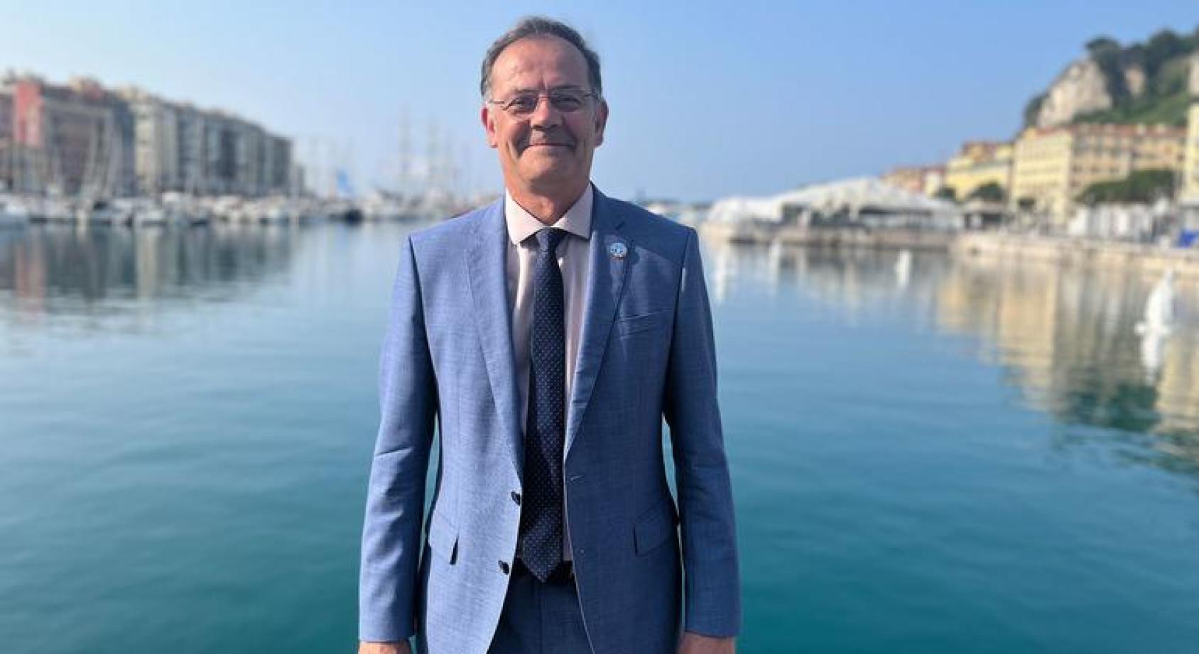 一位联合国官员穿着西装站在蓝色的湖面前面，背影两侧依稀是一些房子