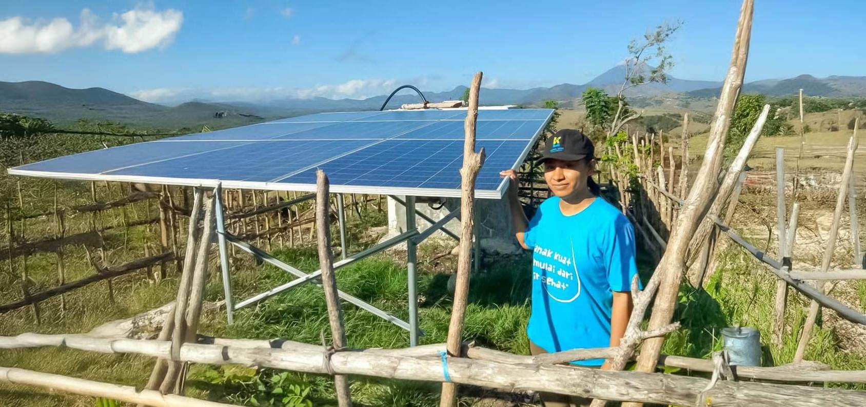 A woman in a blue shirt stands next to a grey solar powered panel in a green field