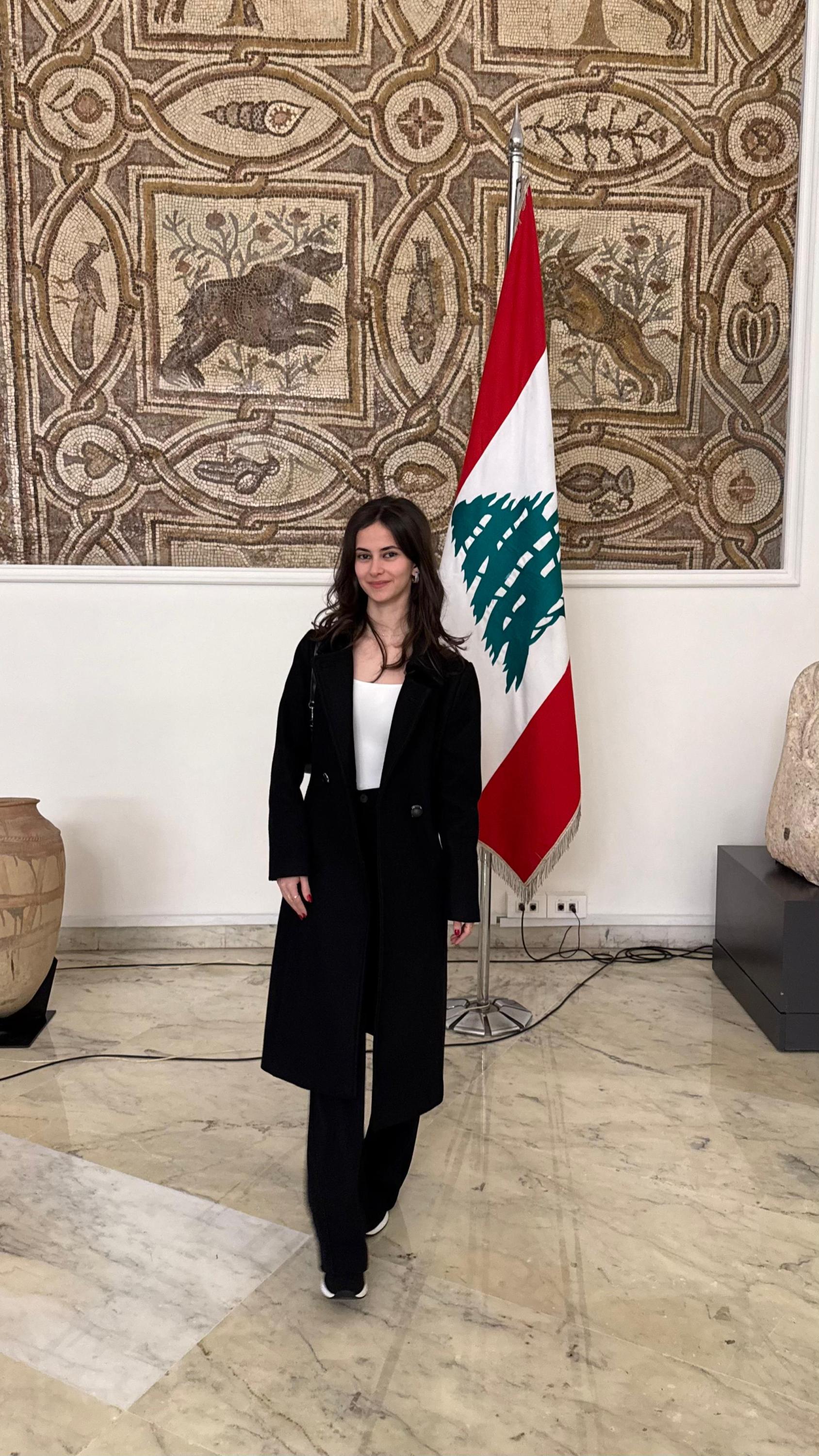 A woman in a dark formal suit stands in front of a flag of Lebanon
