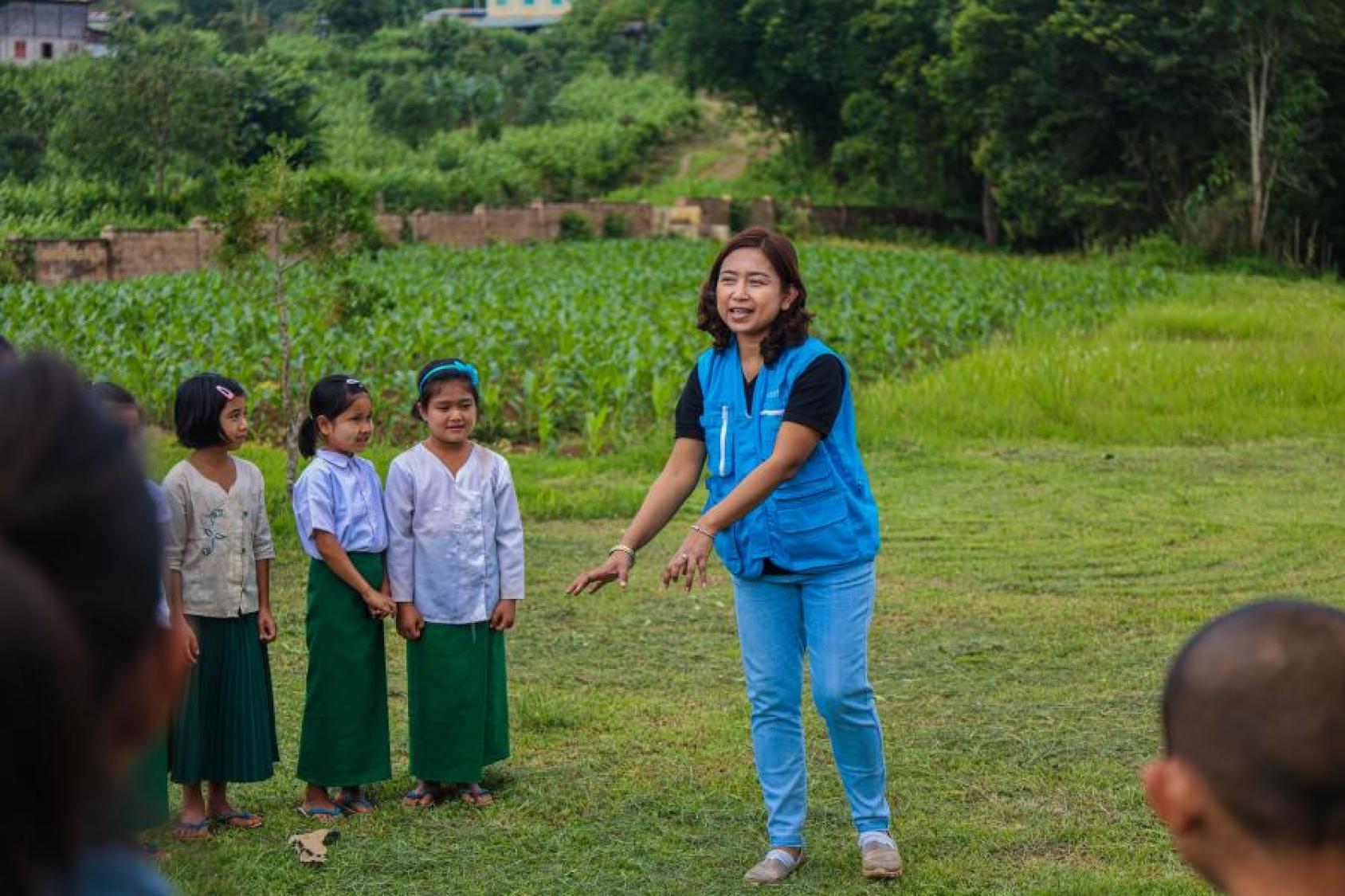 A woman in a blue uniform talks to school students in an outdoor location filled with greenery.