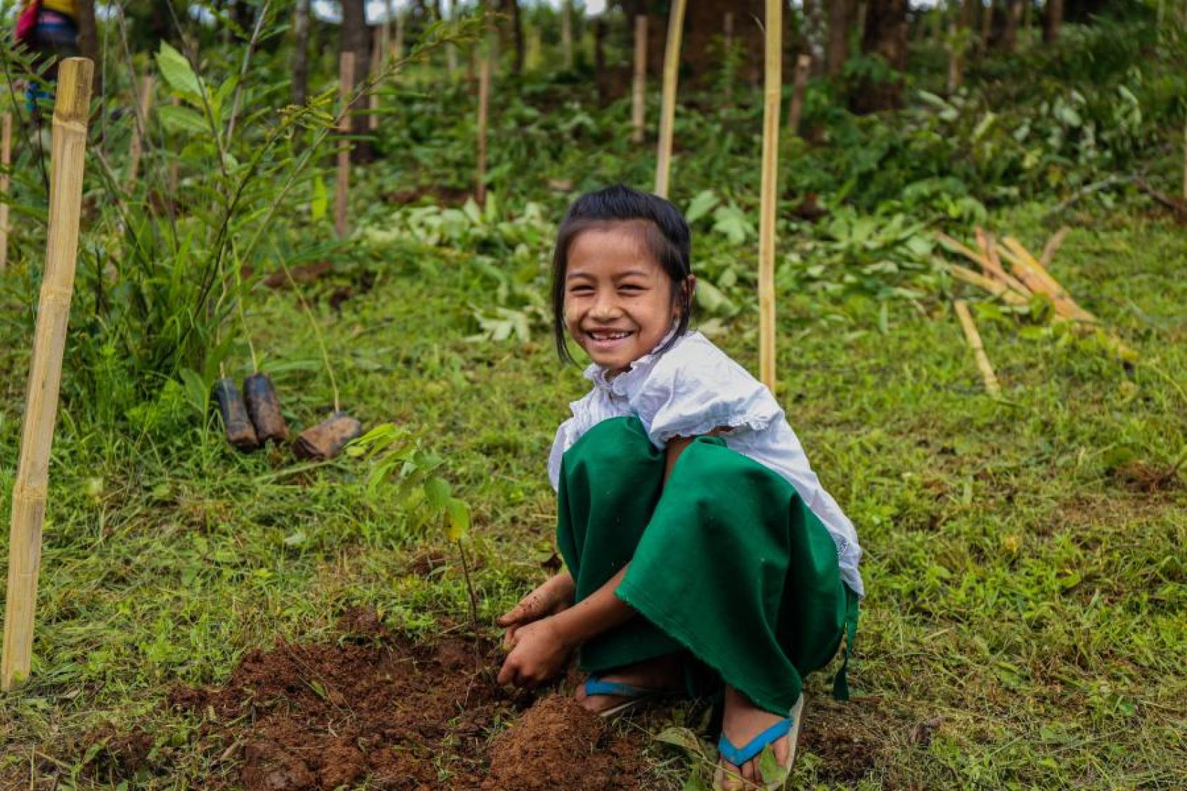 A young girl in white shirt and green pants pats mud on a sapling.