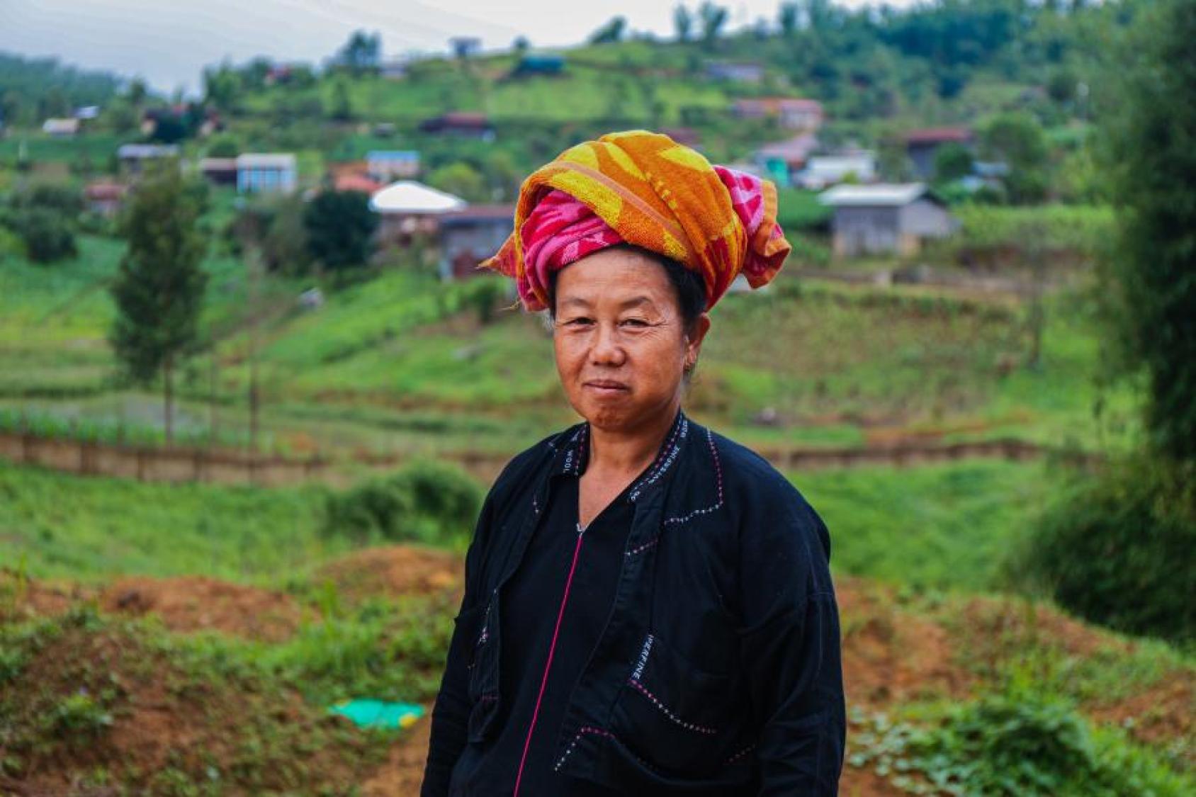 A woman in a a red turban and a black dress stands against a lush green background.