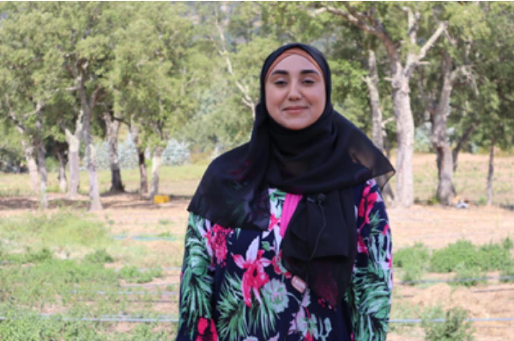 Une femme portant un foulard noir et une robe colorée se tient devant un bosquet d'arbres verts.