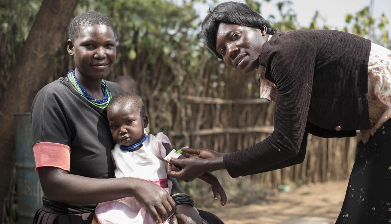 Une mère est assise avec sa petite fille tandis qu'une femme se penche sur fillette pour lui passer une petite bande autour du bras. Toutes trois regardent la caméra.
