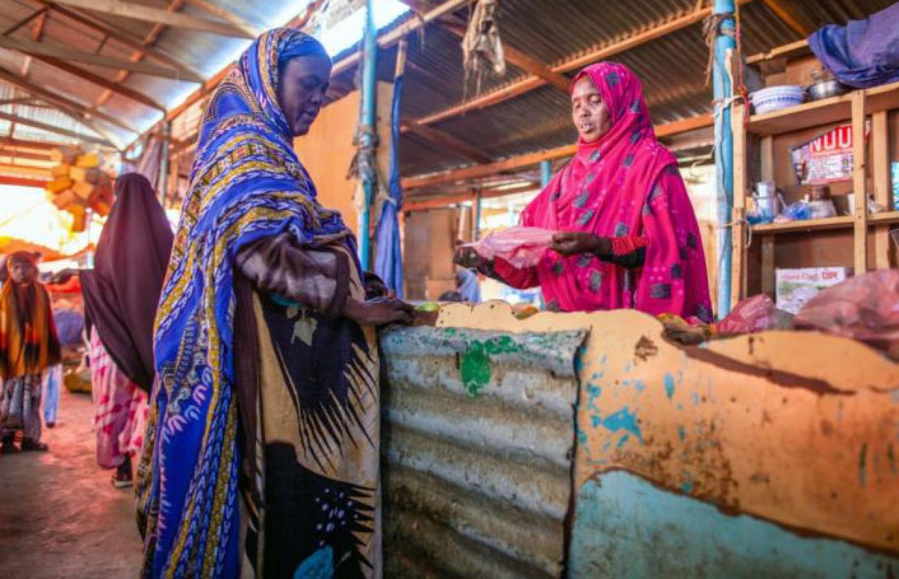 El propietario de un puesto atiende a un cliente. Ambas mujeres llevan vestidos de colores. Otras personas recorren los pasillos del mercado cubierto.