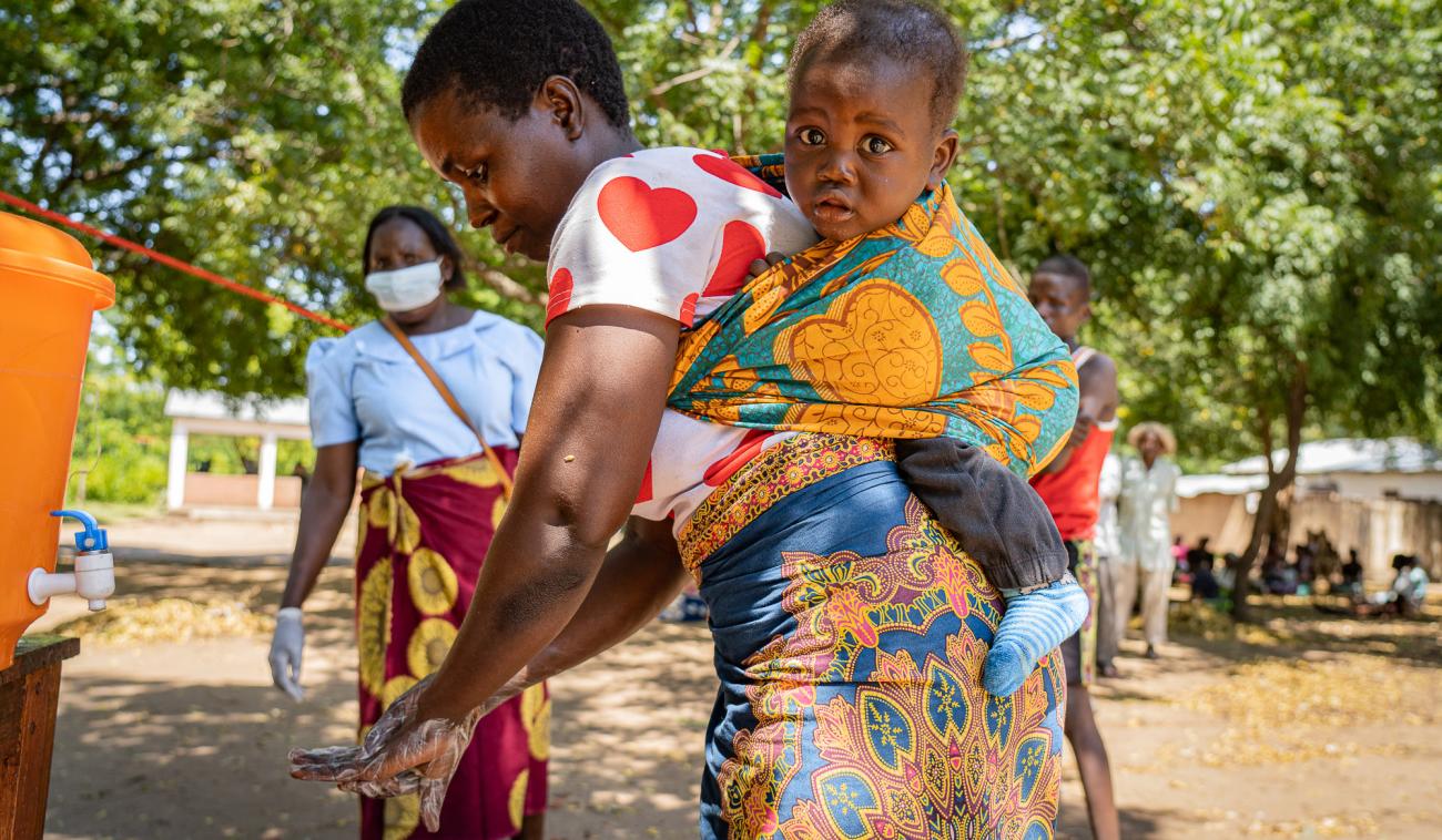 Mother washes her hands while her baby strapped to her back looks at the camera.