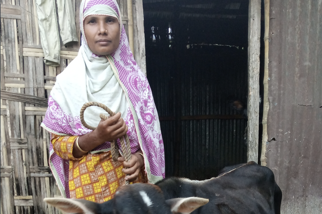 Une femme portant un voile et tenant une vache à l'aide d'une corde se tient debout, face à la caméra, devant une porte en bois.