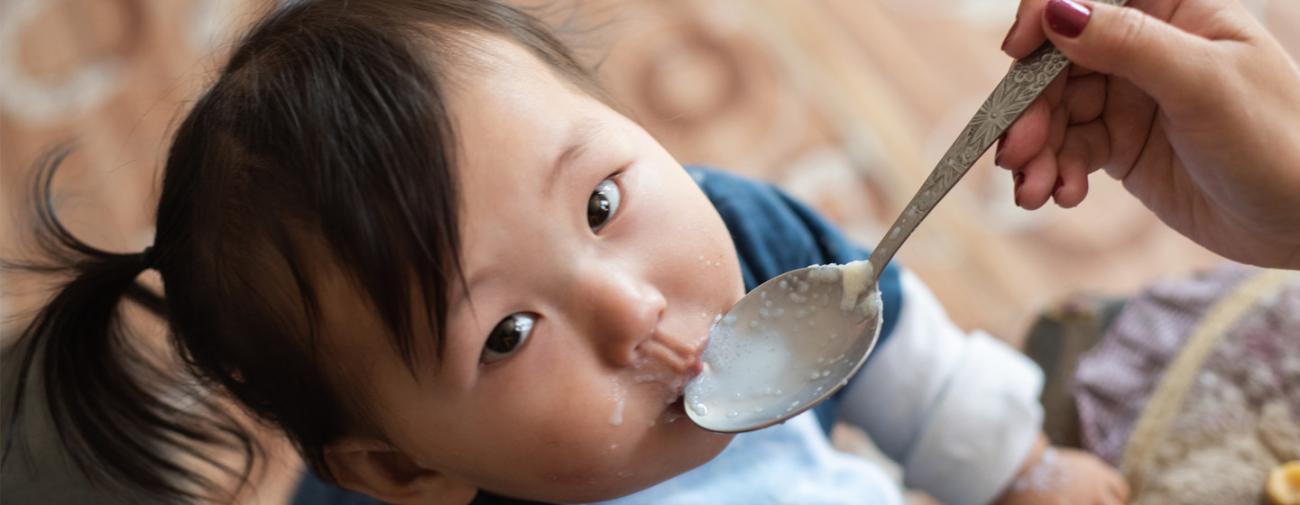 Una niña pequeña mira a la cámara, mientras alguien le da de comer con una cuchara.