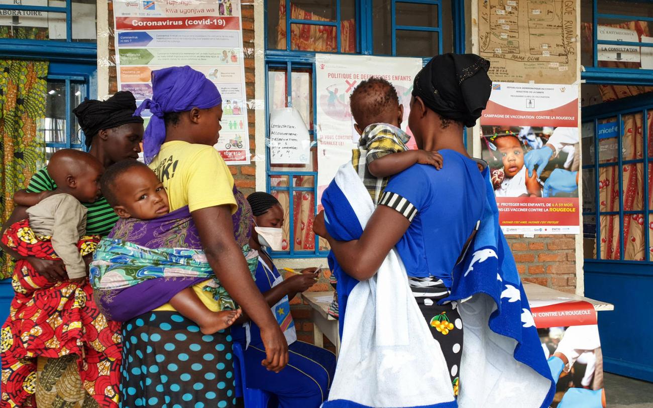 Tres mujeres, cada una con un bebé, se paran frente a la puerta de un centro médico y preguntan con una empleada sentada en una mesa fuera del centro. En la pared exterior del centro se colocaron varios carteles con información sobre COVID-19 y vacunación contra otras enfermedades infecciosas.