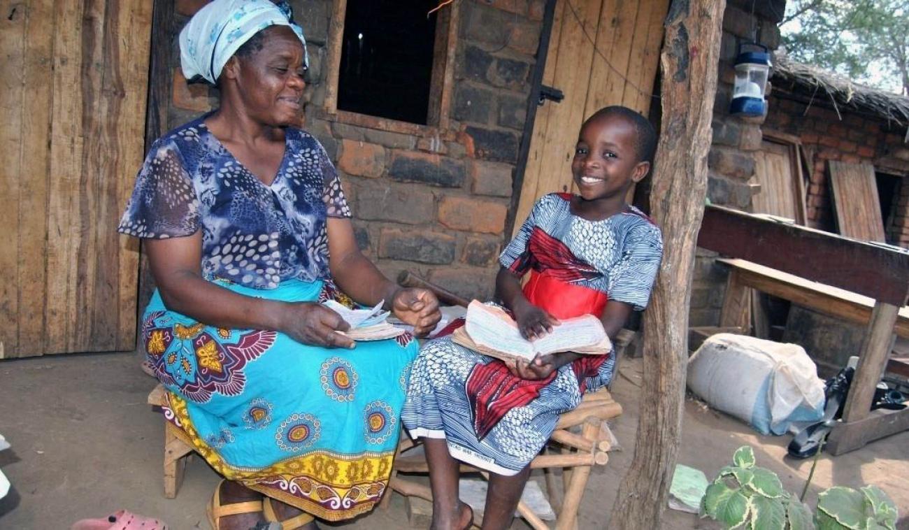Florence hace su trabajo escolar en casa. Su madre siempre la apoya y trata de ayudarla. Florence y su mamá se sientan una al lado de la otra en sillas de madera, frente a una casa hecha con presuntos materiales locales (piedra y madera). La pequeña Florence, a la derecha, tiene la espalda apoyada en un poste de madera. Ella sostiene un cuaderno en sus manos y mira a la cámara con una gran sonrisa. Su madre, a la izquierda, sostiene hojas de papel y mira en dirección a su hija, sonriendo con orgullo.
