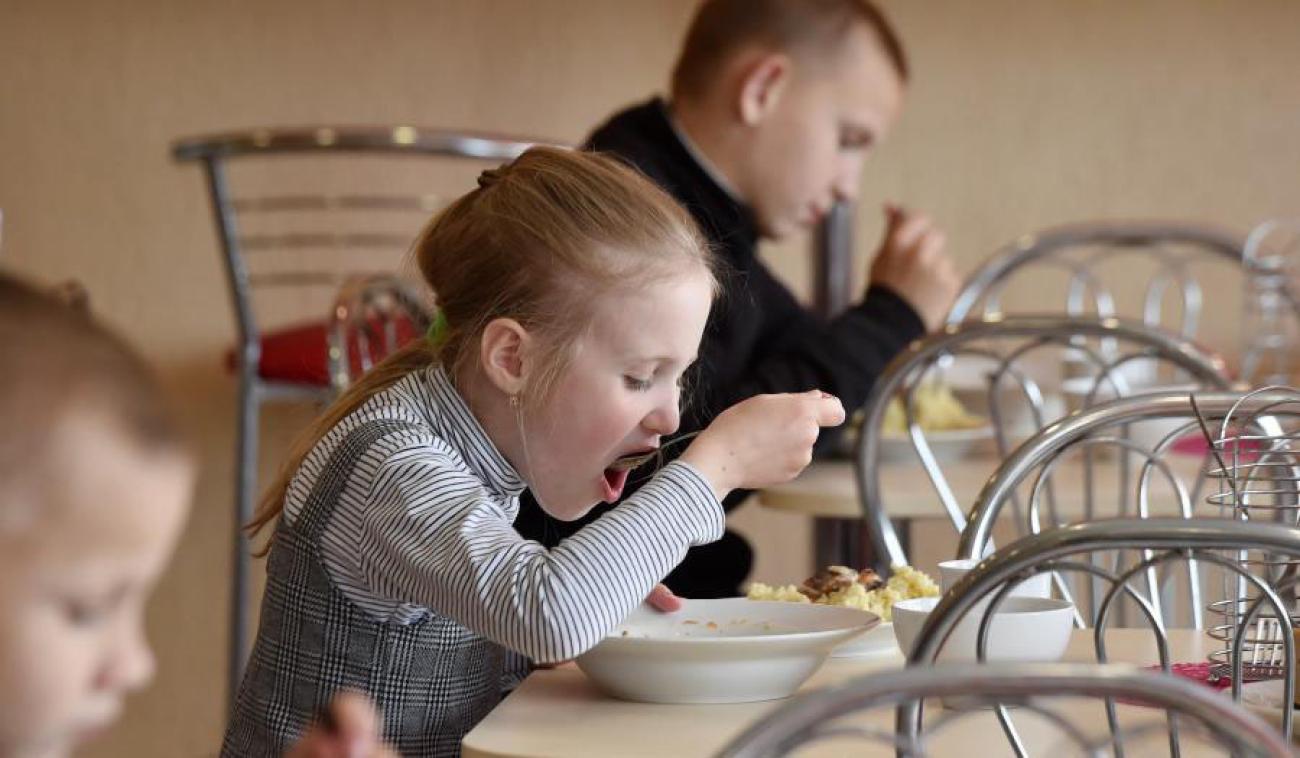 Tres pequeños en edad escolar (una niña y dos niños) comen, cada uno en su propia mesa.
