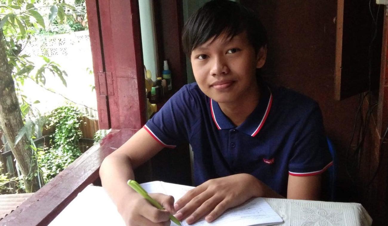 Thaw Lay, un estudiante de Yangon, Myanmar, hace sus deberes en casa. Está sentado a una mesa, cerca de una ventana detrás de la cual se adivina un jardín. Sonríe mientras mira a la cámara.