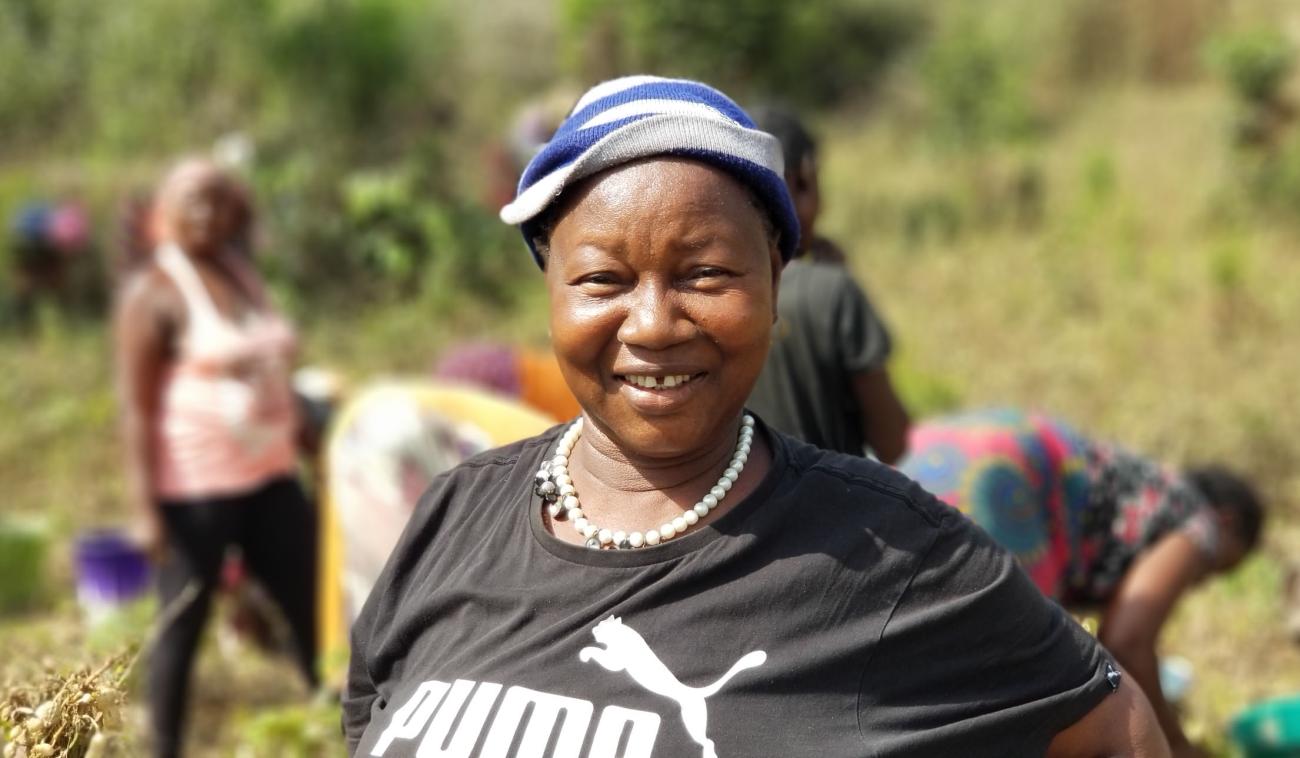 A woman farmer smiles cheerfully at the camera as other women harvest crops behind her.