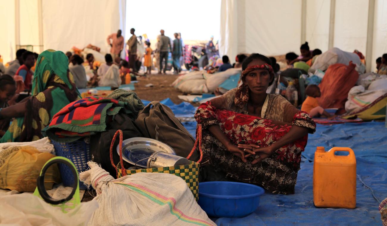 Several people sitting in an indoors market with baskets and a gas can.