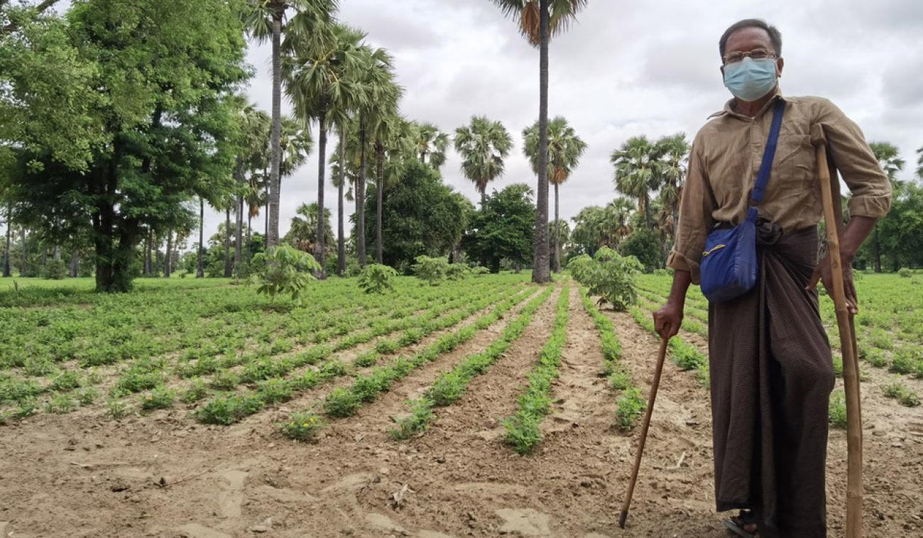 A farmer stands with crutches next to a lush farmland. 