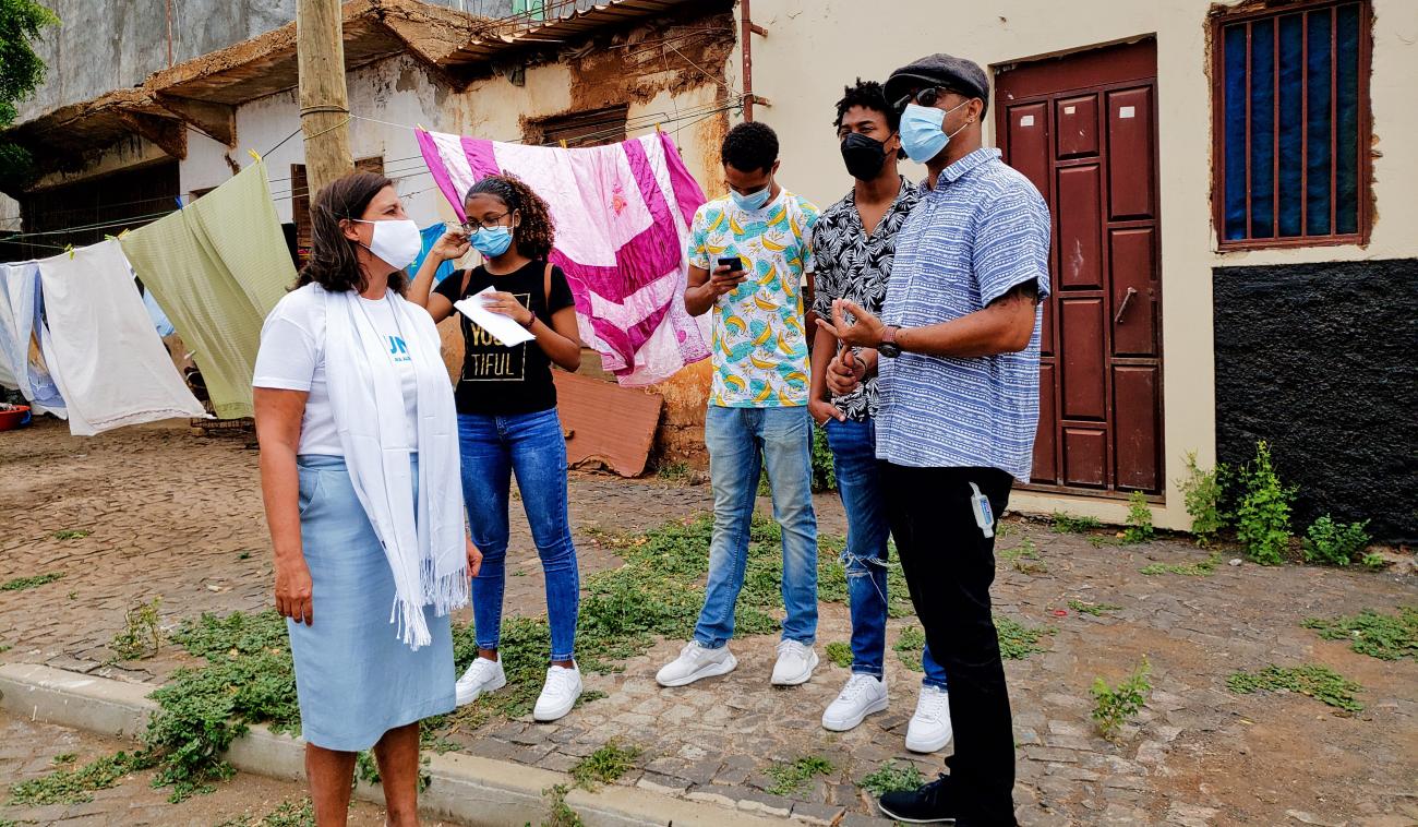 La Coordinadora Residente, Ana Patricia Graça, habla con jóvenes artistas en un semi círculo, frente a las puertas de las casas de una comunidad. Todos usan mascarillas.