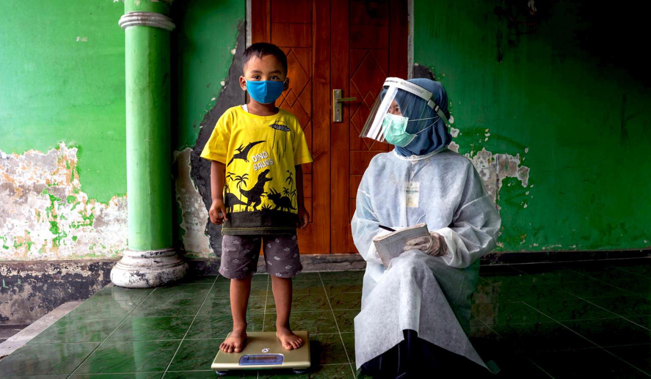 A little boy stands on a scale as he looks at the camera wearing a face mask and a healthcare worker in full head-to-toe PPE kneels by him.