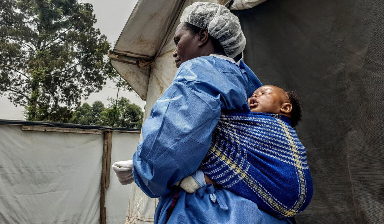 Une femme portant une tenue de protection porte un petit bébé sur son dos.