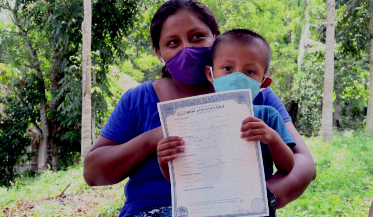Una madre con una mascarilla protectora mira a su izquierda mientras abraza a su hijo. El hijo también usa una mascarilla protectora. El pequeño sostiene con orgullo su certificado de nacimiento, mirando a la cámara.