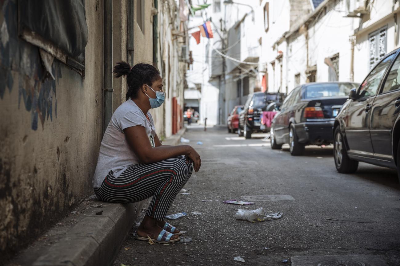 A woman in a blue face mask sits on the side of the road.