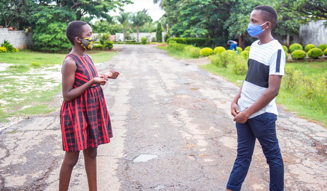 يقف فتى وفتاة يلبسان قناعي وجه متباعدان اجتماعيًا أثناء التحدث في منتصف الطريق في لوساكا، زامبيا.