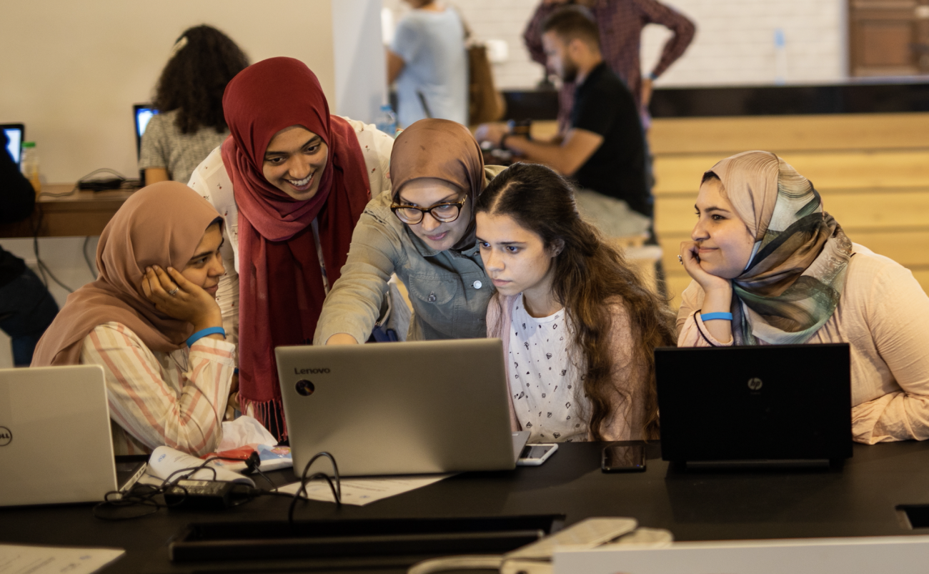 5 girls look at a laptop together in a room with several other people.