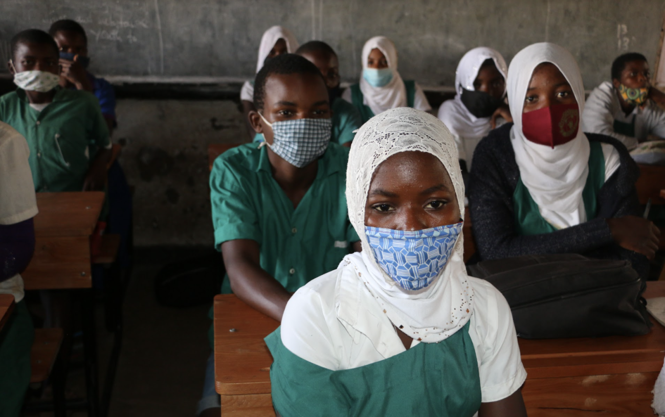 Un grupo de niños y niñas con uniformes escolares y mascarillas se sientan en un aula de frente a la cámara.