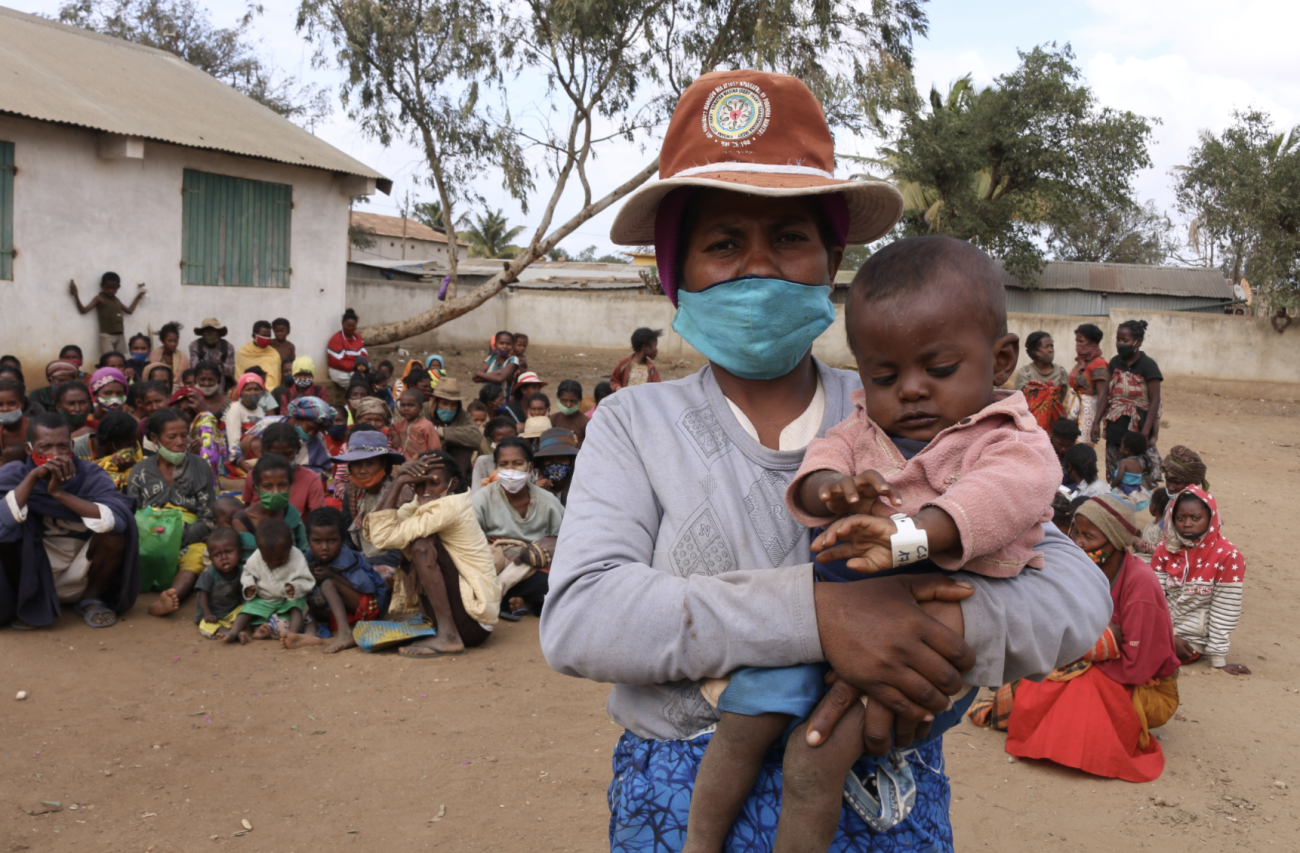 A woman in a mask and a hat holds a baby in her arms while a large group of people sit behind her.
