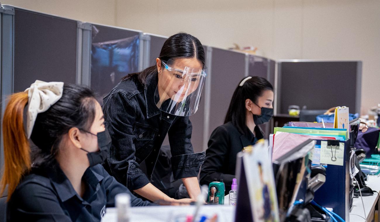 Mujeres con mascarillas y protectores faciales trabajan juntas en un escritorio de una oficina.