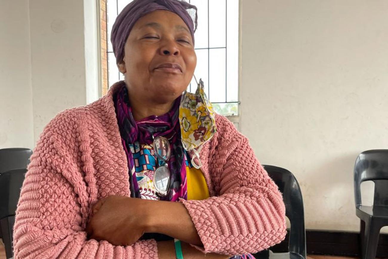 一位穿着粉红色毛衣的老年妇女双臂交疊坐在一个白色的房间里。