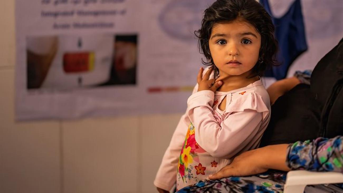 A little girl in a pink shirt looks directly at the camera while she leans on a woman.