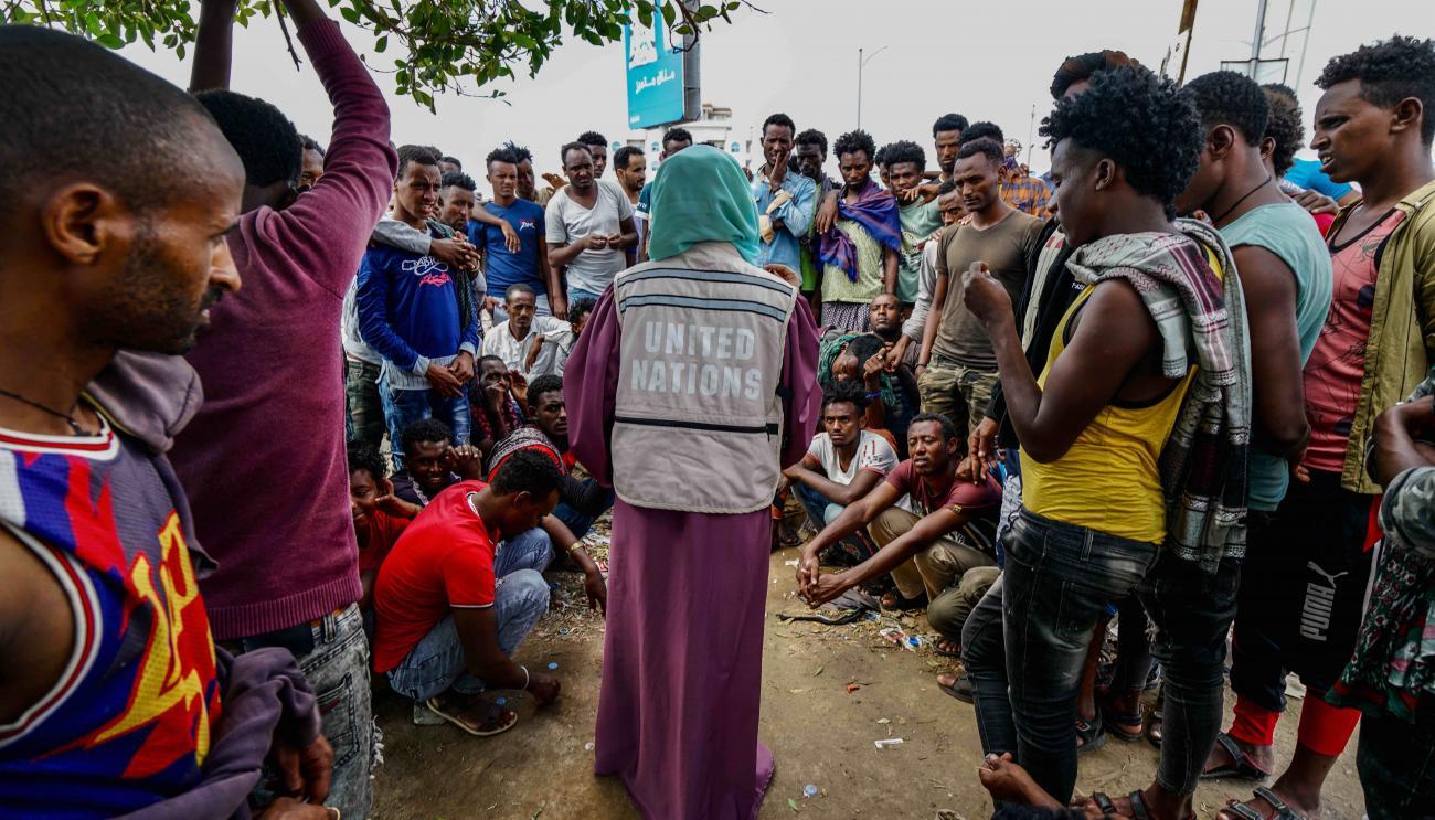 A woman in a United Nations vest is surrounded by many people waiting to receive humanitarian assistance.