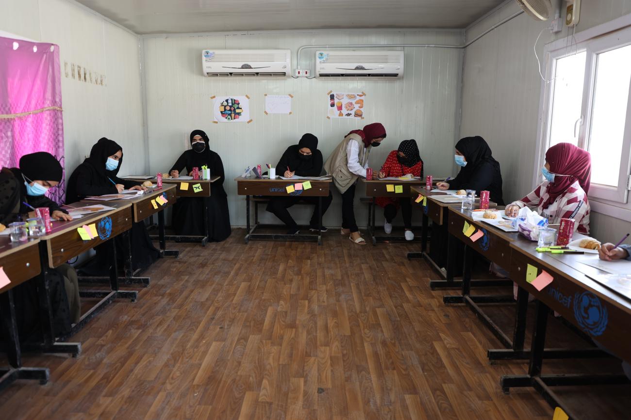 A group of women work at desks with an instructor helping one student. 