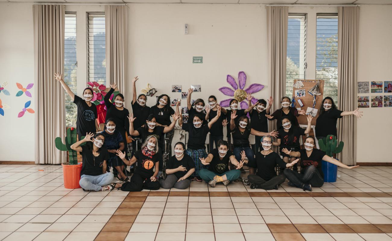 Members of the organization "Mujeres con Capacidad de Soñar a Colores" pose together against a wall as they cheerfully spread their arms towards the front.