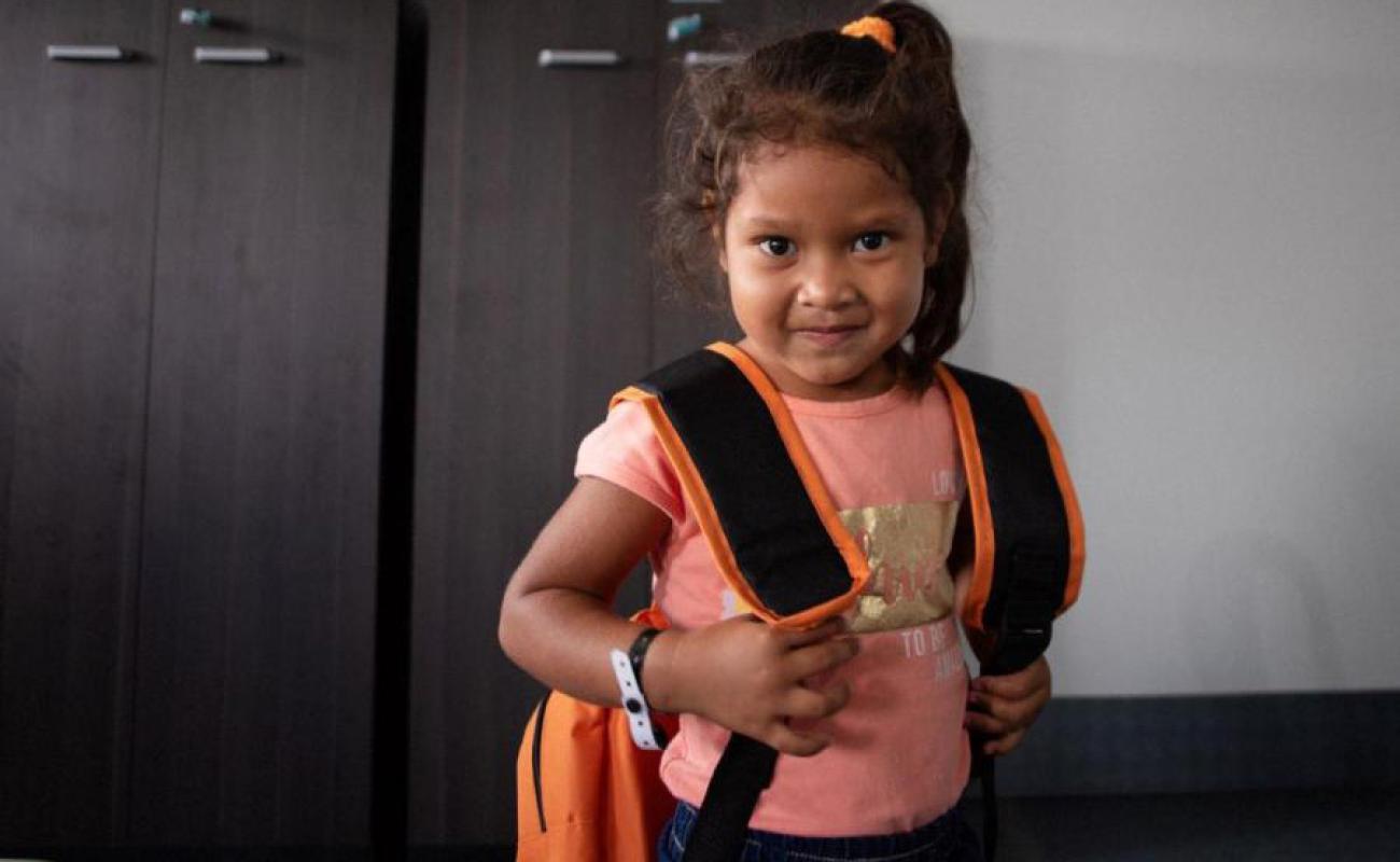 A little girl with a small smile and looks stoically towards the camera as she grasps the straps of her backpack.